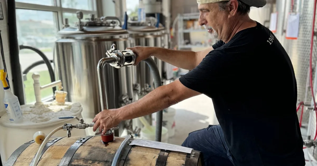 Brewer working at a barrel in the Swan Brewing production space, fermentation tanks and large windows behind him